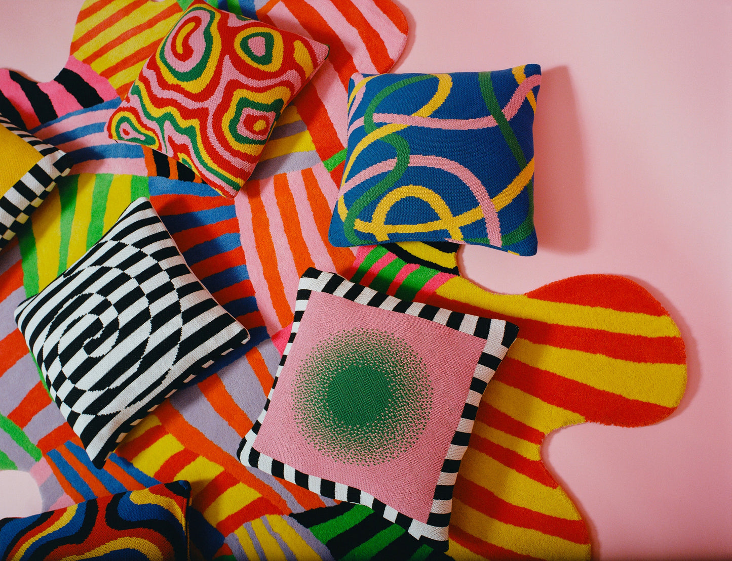 Various knit pillow covers laying on top of the squiggle stripe rug against a pink floor.
