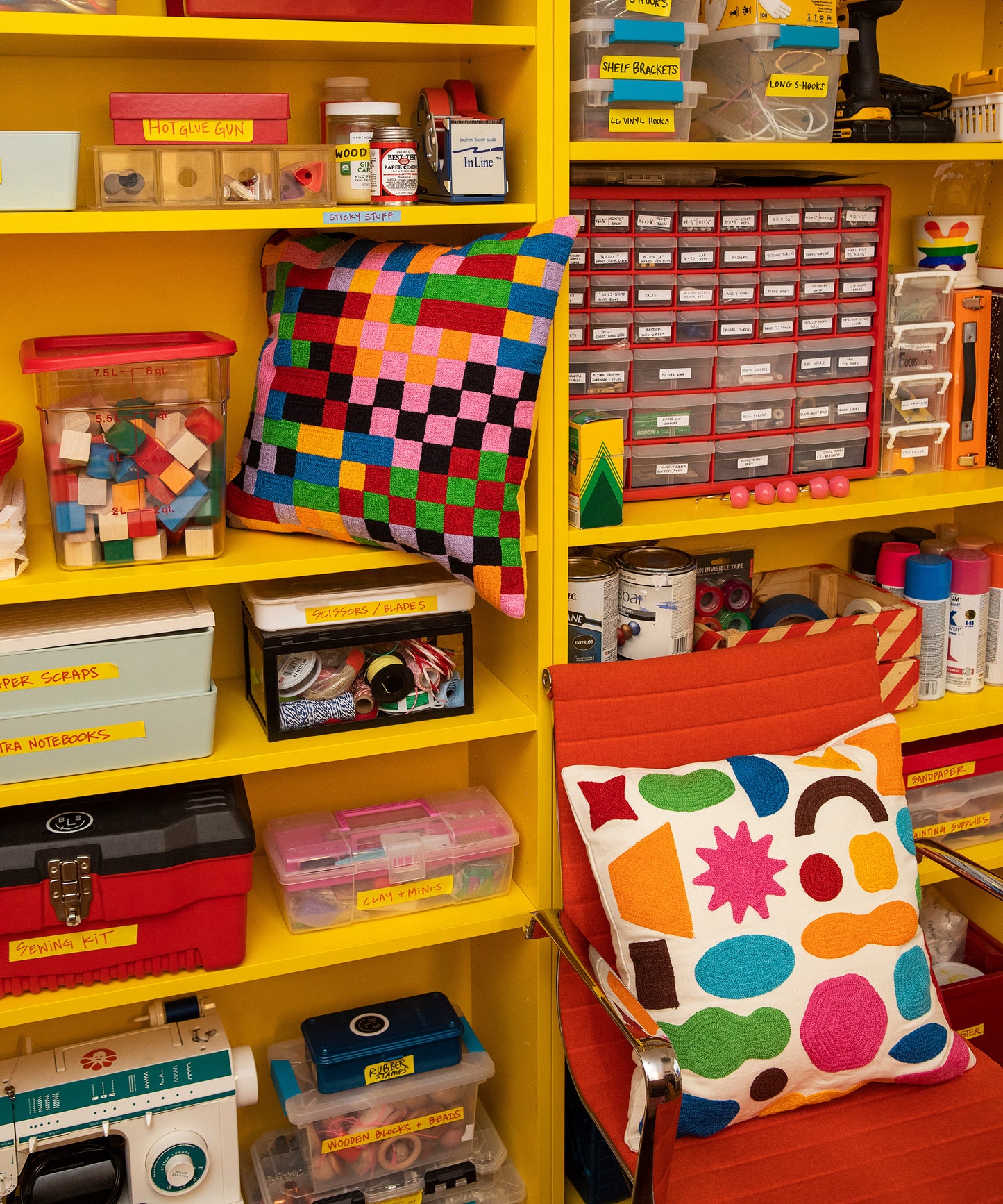 Detail of Pixels pillow sitting in shelf on a yellow bookcase with crafts and the Odds and Ends pillow to the right sitting on top of an orange swivel chair.