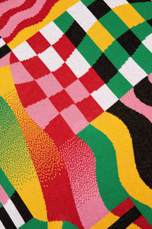 Close up of the Gradient Stripe Blanket showing pink, red and white checkerboard next to a yellow, green, black and white checkerboard surrounded by colored wiggly stripes.
