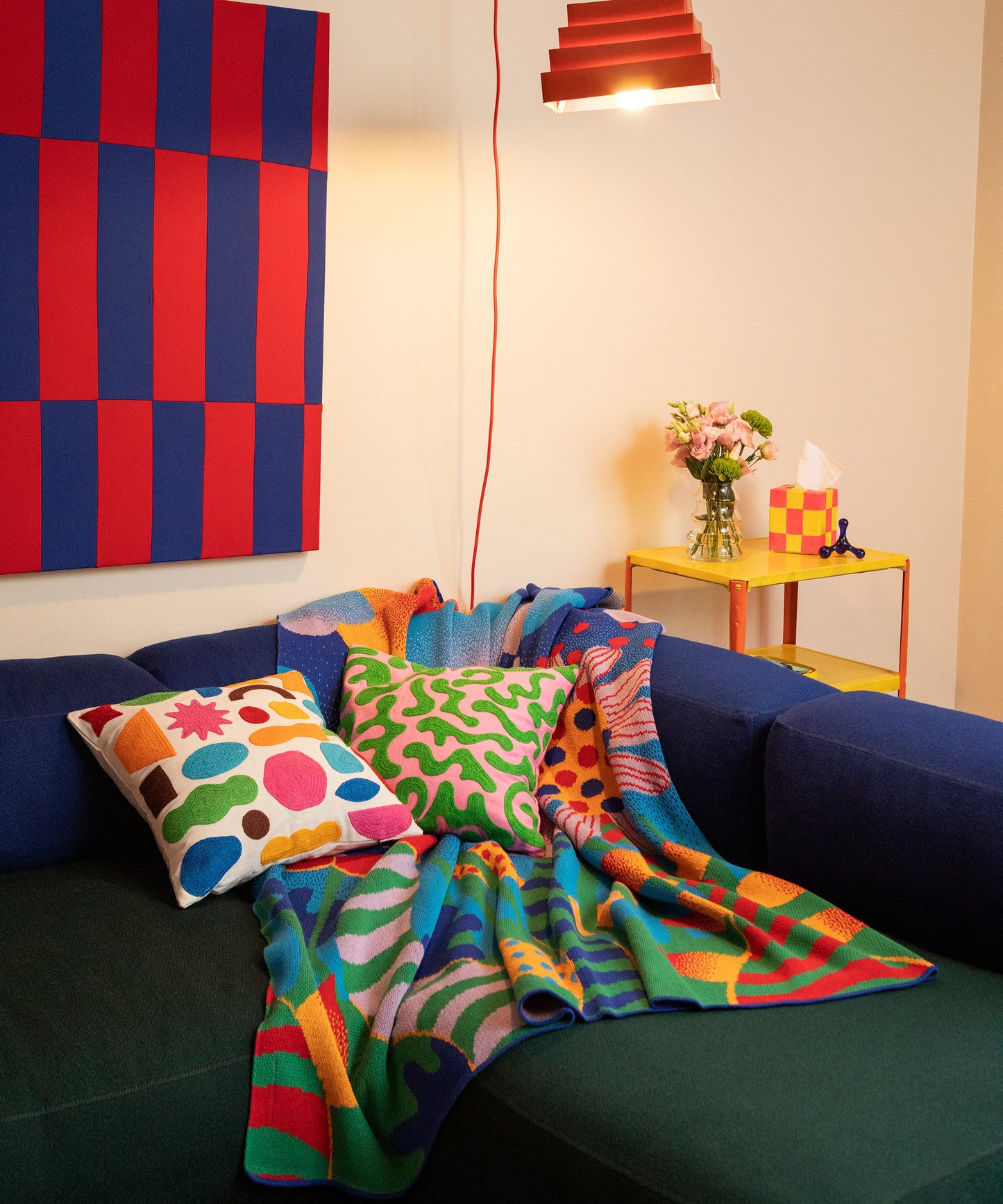 Image of the Best Buds Blanket sprawled over a blue and green sofa located in a colorful living room. On top of the blanket sits the Odds and Ends Pillow Cover and Silly Squiggles Pillow Cover. 