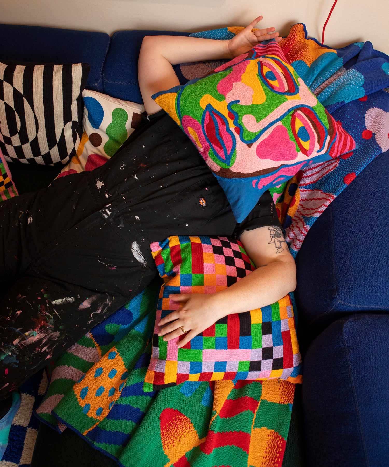 Image of a person laying on top of the Best Buds Blanket that is sprawled over a blue and green sofa. This person has the Portrait Pillow Cover resting on their face with the Pixels Pillow Cover under their left arm. 