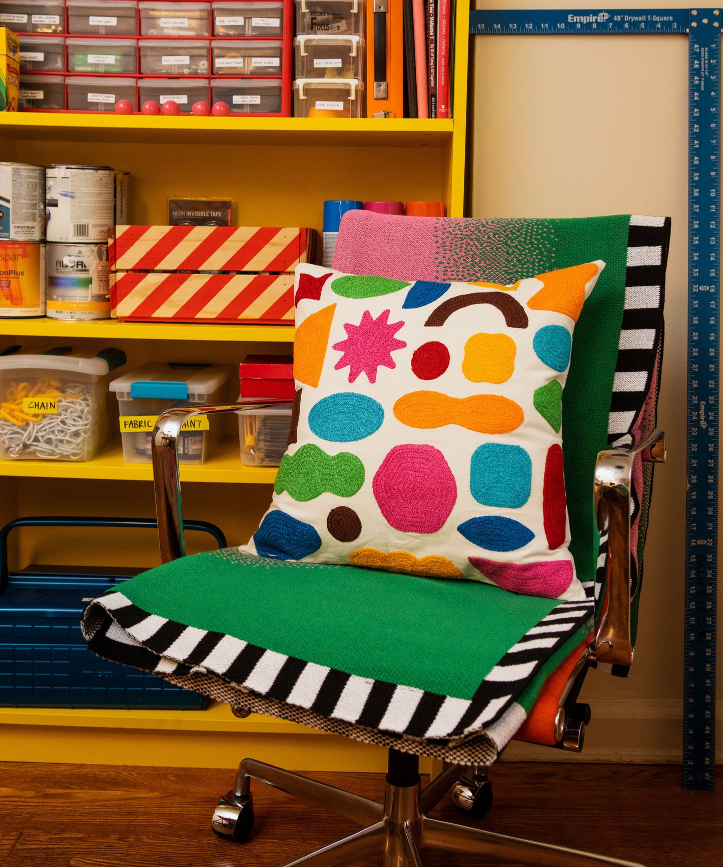 Detail image of Burst Blanket folded vertically on a swivel chair that stands in front of a bookshelf filled with supplies. On top of the blanket sits the Odds and Ends Pillow Cover. 