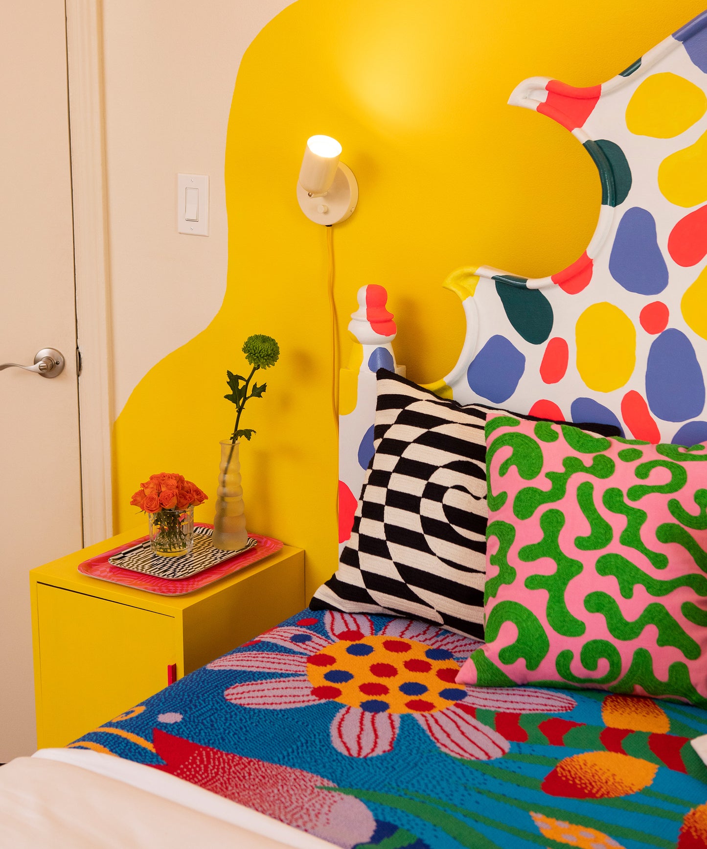 Detail of a bed with the Best Buds Blanket and the Silly Squiggles Pillow sitting on top of it. Sitting on the left is the Dazzle Pillow and to the left is a yellow nightstand with the Wiggles and Waves and Dazzle Trays stacked.