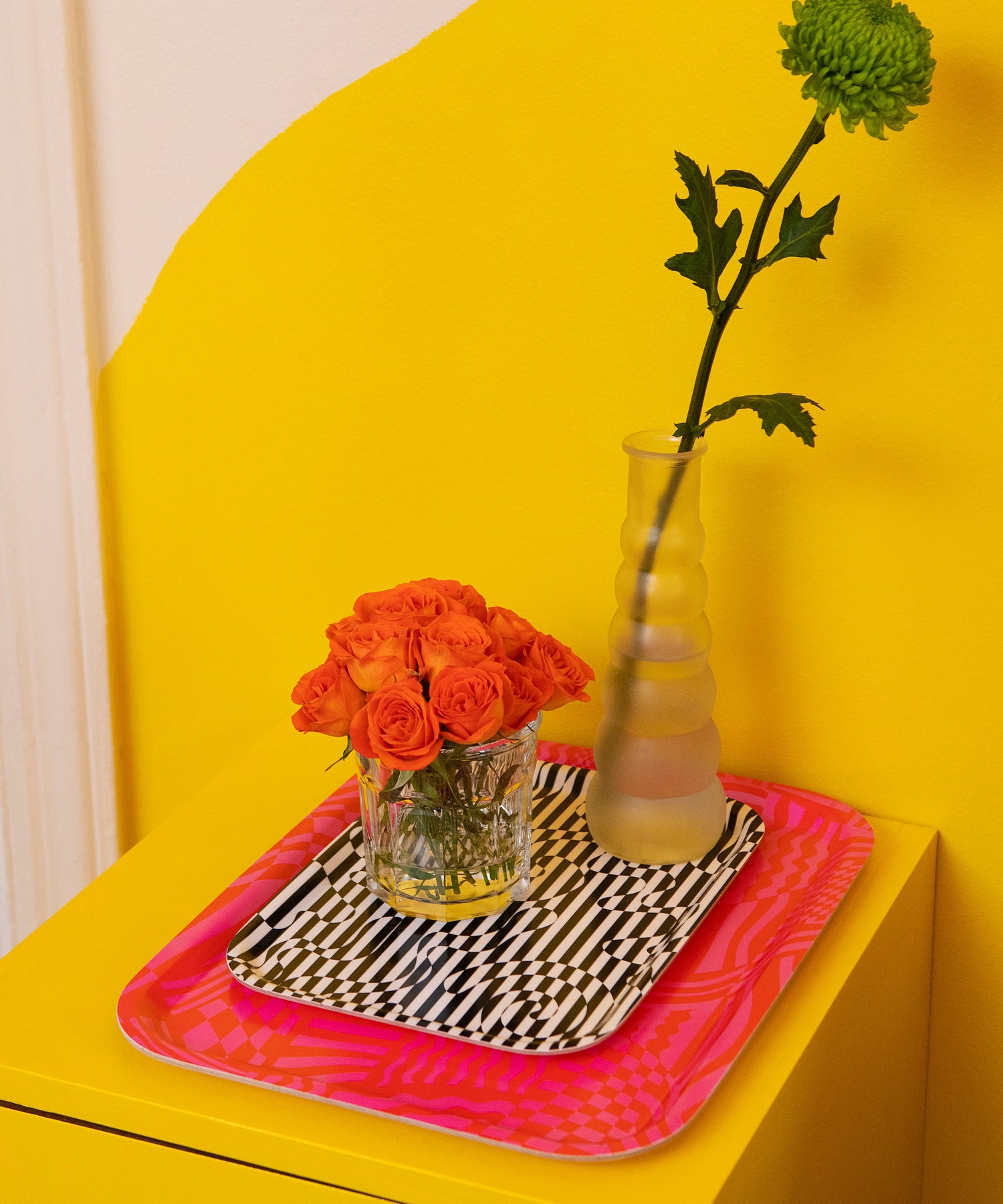 Detail of the Dazzle Tray stacked on top of the Wiggles and Waves Tray holding a small and large flower vase. These are sitting on top of a yellow nightstand.